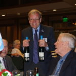 An older man holding up two bottles of white, smiling.