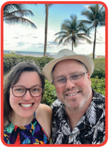 A woman wearing glasses and a man wearing glasses and a hat, dressed in tropical outfits smiling in front of beach with palm trees behind them.