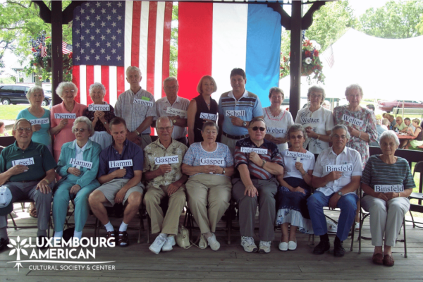 Group of people posing in front of a Luxembourg and American Flag each holding a small sign with a last name on it