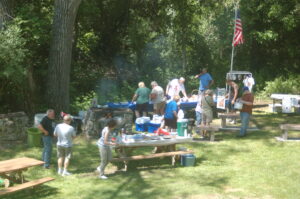People sitting on picinic tables eating a picinic in area shaded by trees.